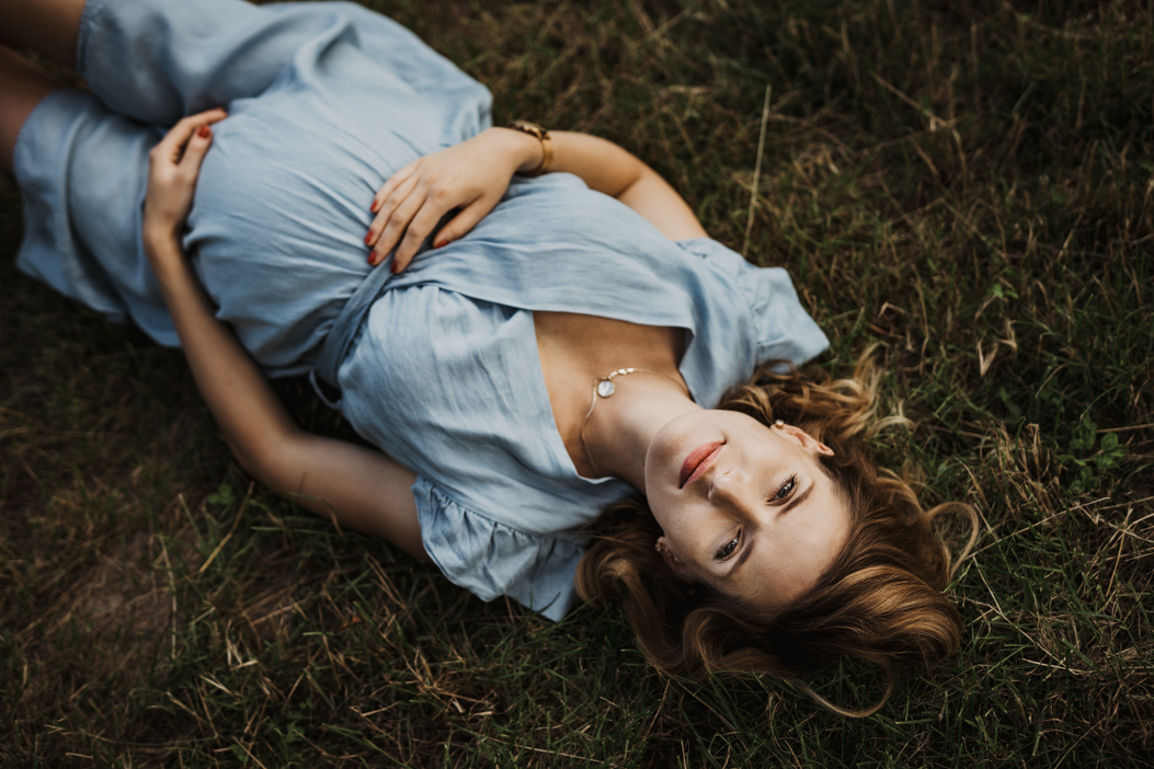 młoda kobieta w ciąży leżąca na trawie i pozująca do zdjęcia na plenerowej sesji ciążowej