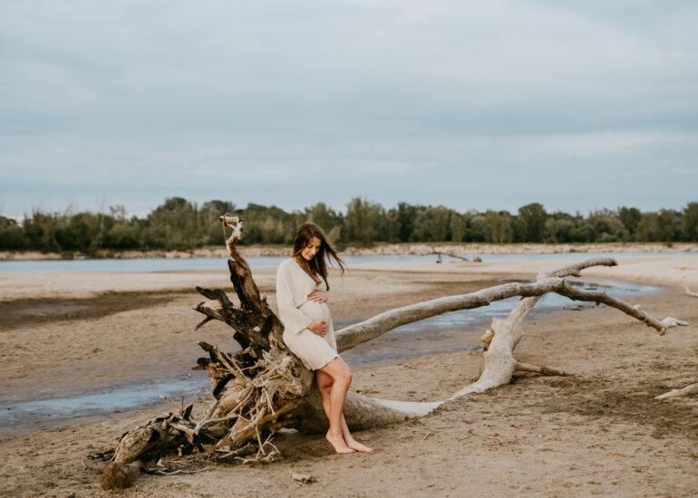 kobieta w ciąży opierająca się o wyrzucony przez wodę na plażę konar drzewa