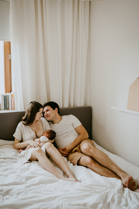rodzice siedzący z noworodkiem na łóżku z białą pościelą na domowej sesji zdjęciowej w warszawie