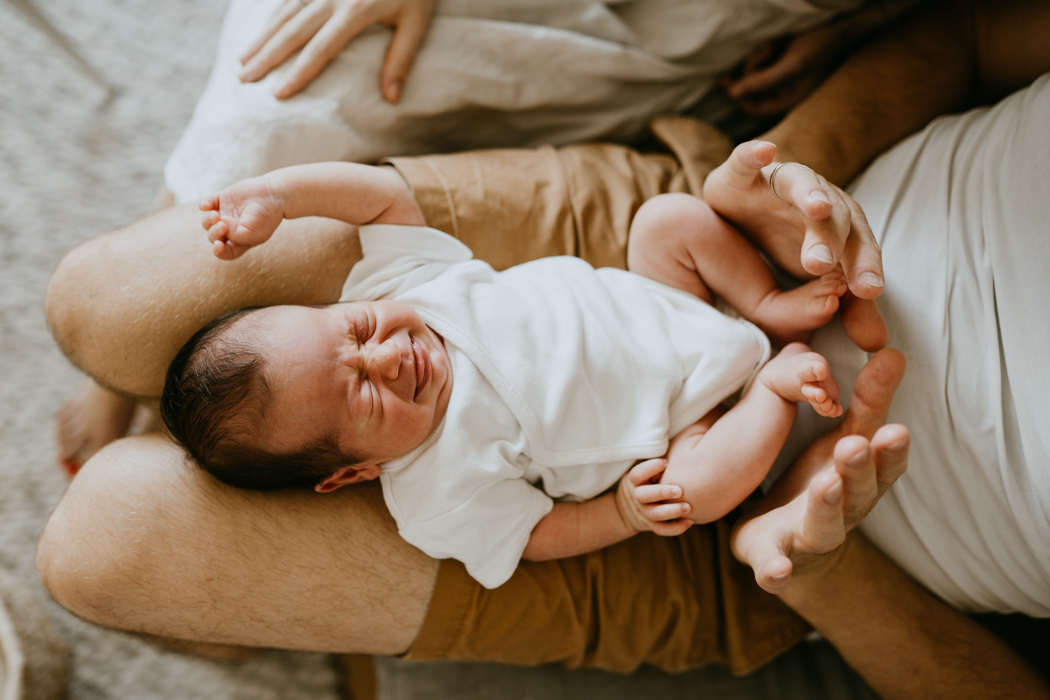 noworodek w białym body ze śmieszną minką leżący u taty na kolanach podczas naturalnej sesji zdjęciowej w warszawie