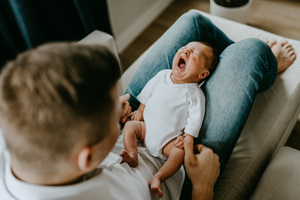 noworodek w białm bodziaku płaczący u taty na kolanach podczas domowej sesji zdjęciowej