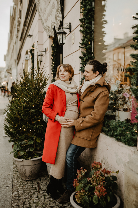 kobieta w ciąży w beżowej opiętej sukience i czerwonym płaszczu pozująca ze swoim mężczyzną do zdjecia na tle świątecznej witryny sklepowej