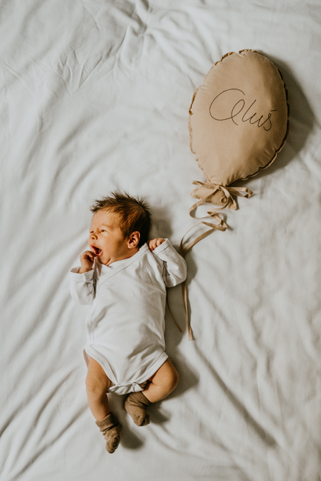 noworodek w białym body i skarpetkach leżący na łóżku z balonkiem ze swoim imieniem