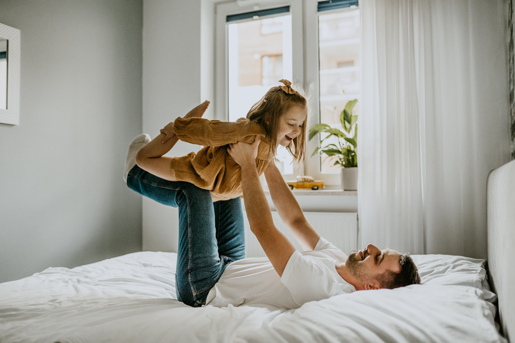 dziewczynka ćwicząca akrobatykę z tatą na łóżku w sypialni podczas domowej sesji rodzinnej