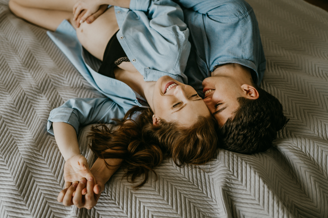 uśmiechnięta para w ciąży ubrana w niebieskie koszule, leżąca na łóżku podczas naturalnej sesji ciążowej