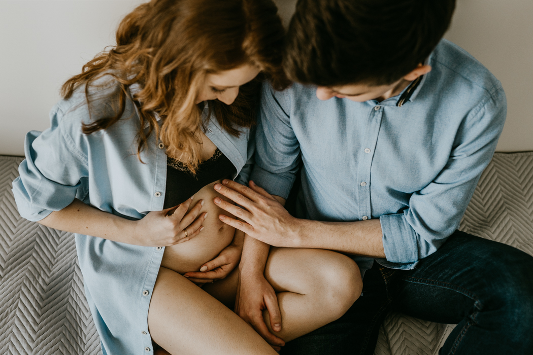 kobieta i mężczyzna siedzący na łóżku wpatrują się i głaszczą brzuch podczas sesji ciążowej w domu w warszawie