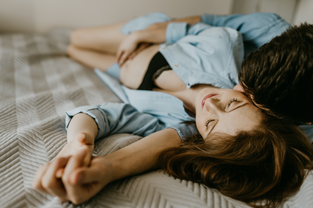 para leżąca i przytulająca się na łóżku ze splecionymi dłoniami podczas domowej sesji ciążowej