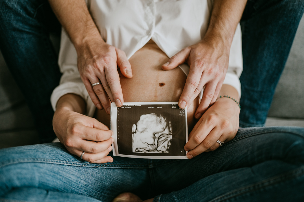 fotografia dłoni pary trzymajacych USG na tle brzuszka ciążowego 