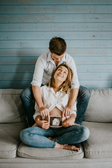zdjęcie przyszłych rodziców siedzących na kanapie i trzymajacych USG na tle brzuszka ciążowego wykonane na domowej sesji ciążowej