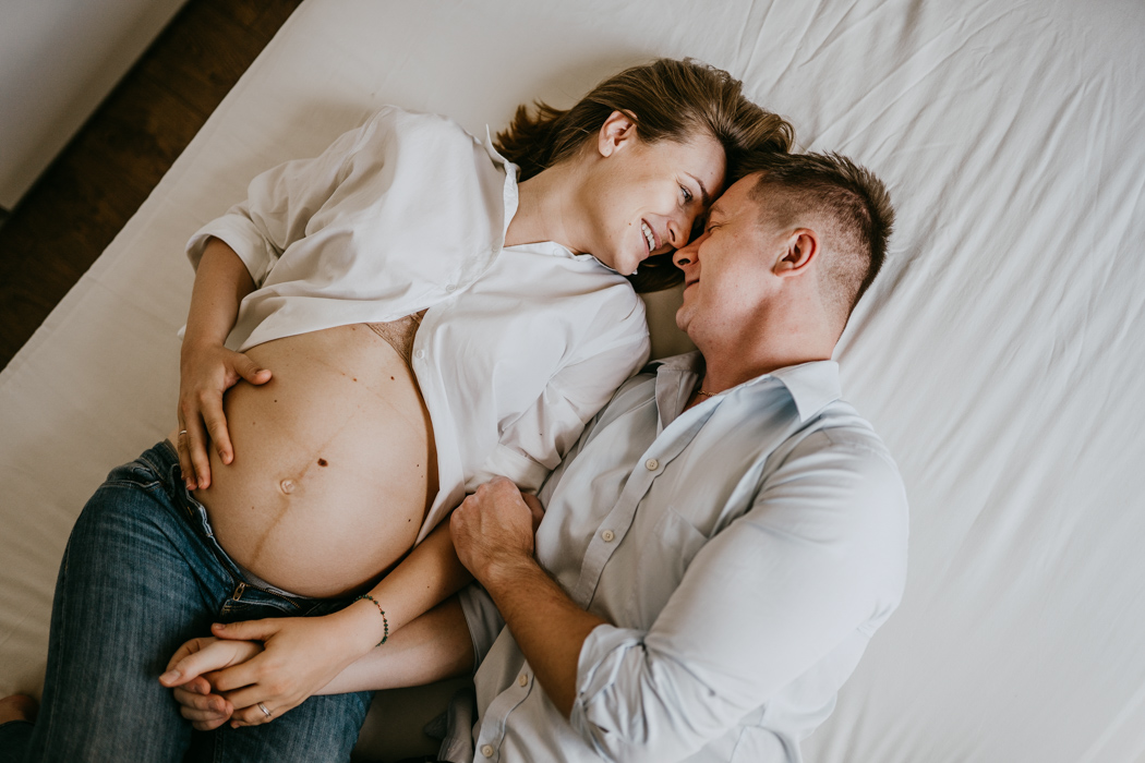 klimatyczne zdjęcie pary w ciąży leżącej na łóżku i śmiejącej się do siebie wykonane na sesji lifestyle w WARSZAWIE