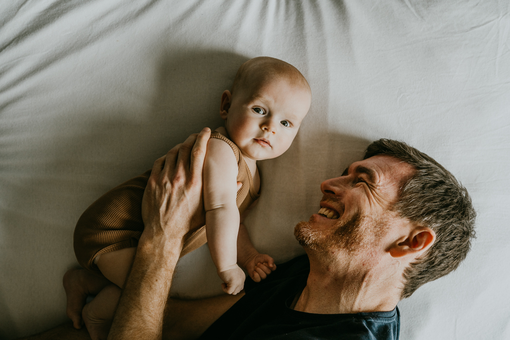 urocze zdjęcie uśmiechniętego taty i niemowlaka na białej pościeli podczas sesji rodzinnej w domu