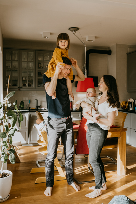 fotografia rodzinna na której mama trzyma niemowlaka na rękach a tata córeczkę na barana