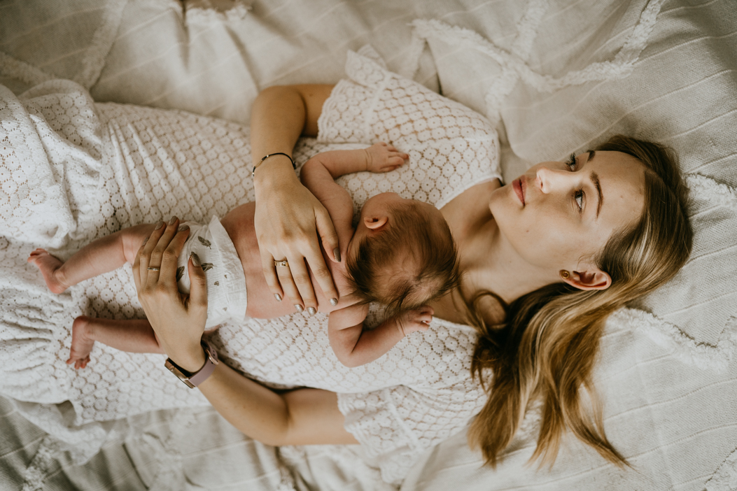 piękna mama leżąca na łóżku z białą pościelą, trzymająca noworodka na rękach