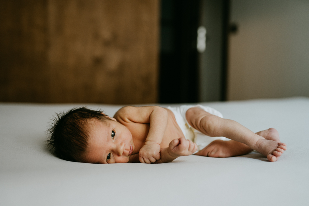 fotografia noworodka z długimi ciemnymi włosami leżącego na łóżku w samym pampersie podczas sesji noworodkowej w domu