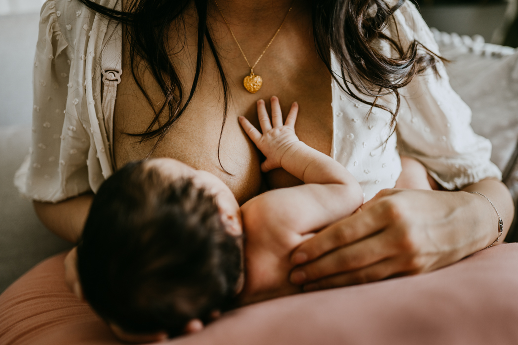 czułe zdjęcie mamy karmiącej noworodka na którym noworodek położył swoją dłoń na klatce piersiowej mamy obok wiszącego wisiorka w kształcie serca
