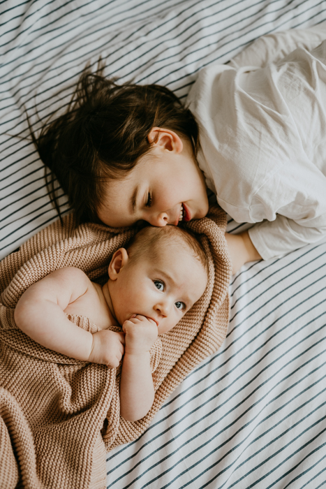 naturalne zdjęcie rodzeństwa leżącego na pościeli w sypialni na którym starszy brat przytula się do niemowlaka podczas sesji noworodkowej w domu w warszawie