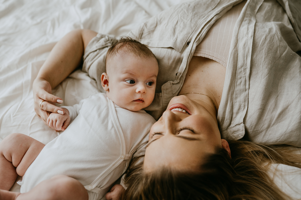 młoda blondwłosa mama leżąca na łóżku w białej pościeli z córeczką podczas sesji niemowlęcej w domu