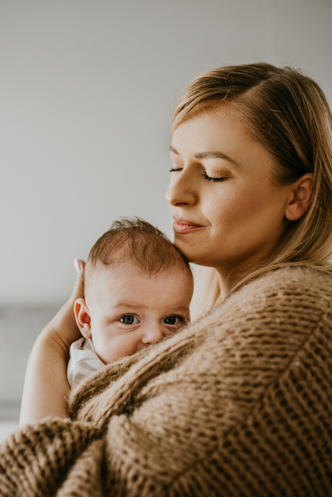 blondwłosa kobieta w karmelowym swetrze z trzymiesięcznym dzieckiem na rękach podczas niemowlęcej sesji w domu
