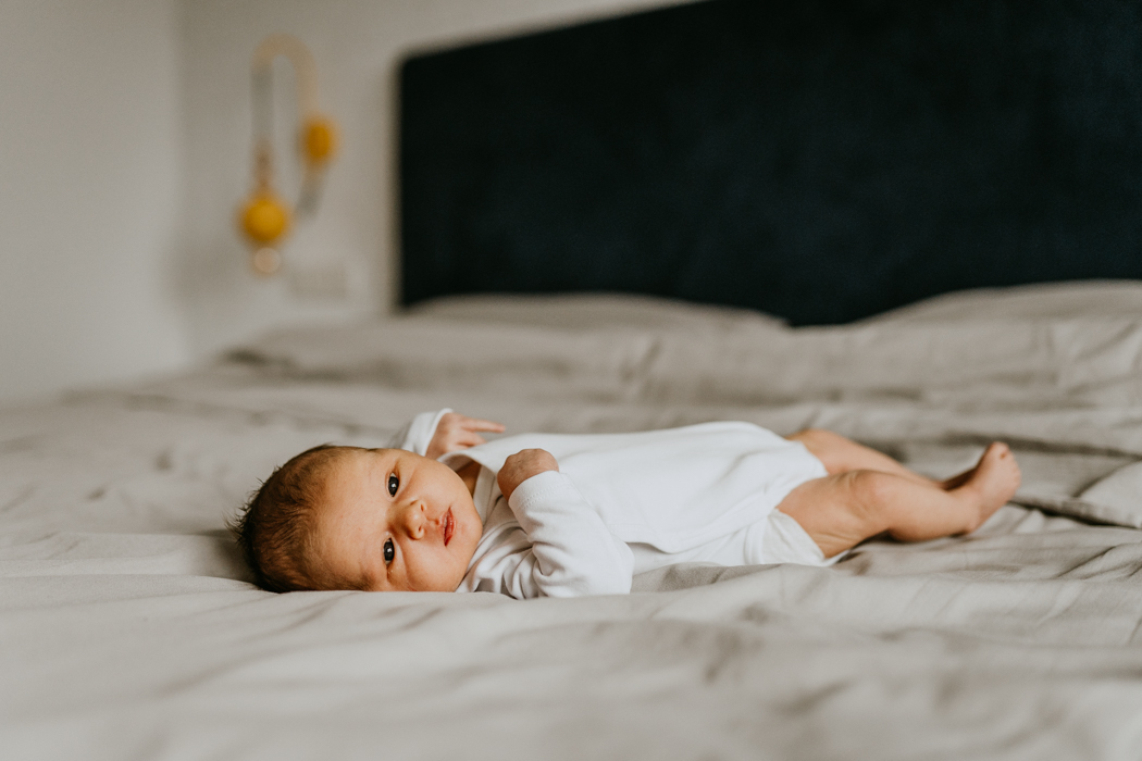 noworodek w białym body leżący w sypialni na szarej jednolitej pościeli podczas sesji zdjęciowej