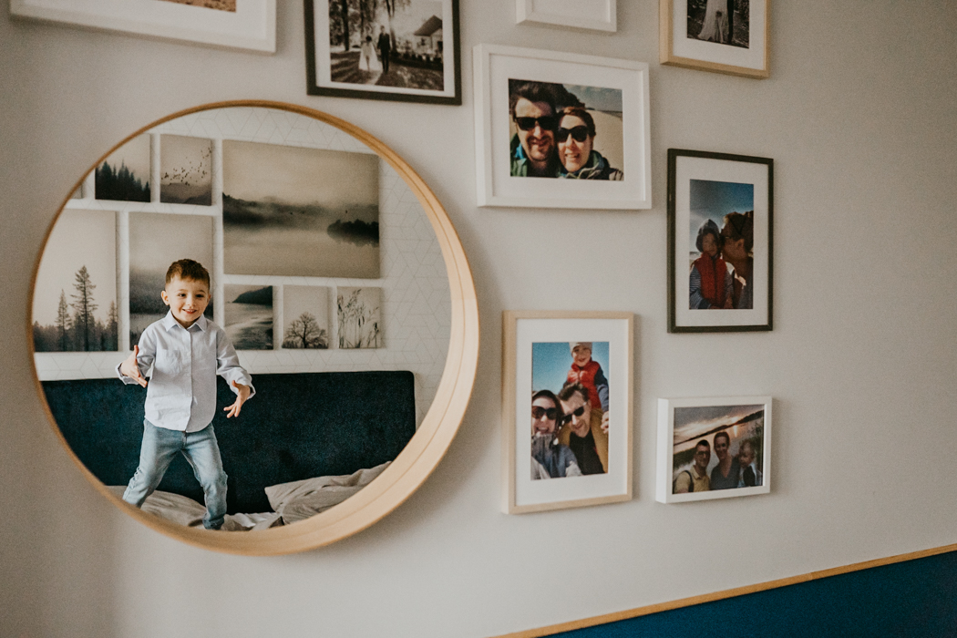 zdjęcie ściany z fotografiami w ramce oraz z lustrem w którym odbija się skaczące na łóżku dziecko